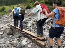 Alpenkamp-deelnemers steken balancerend over een balk een rotsig riviertje over
