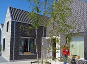 Een hoekje van één van de woningen in Varsenare, met op de voorgrond een jonge boom; langs de huisgevel wandelt een bewoner